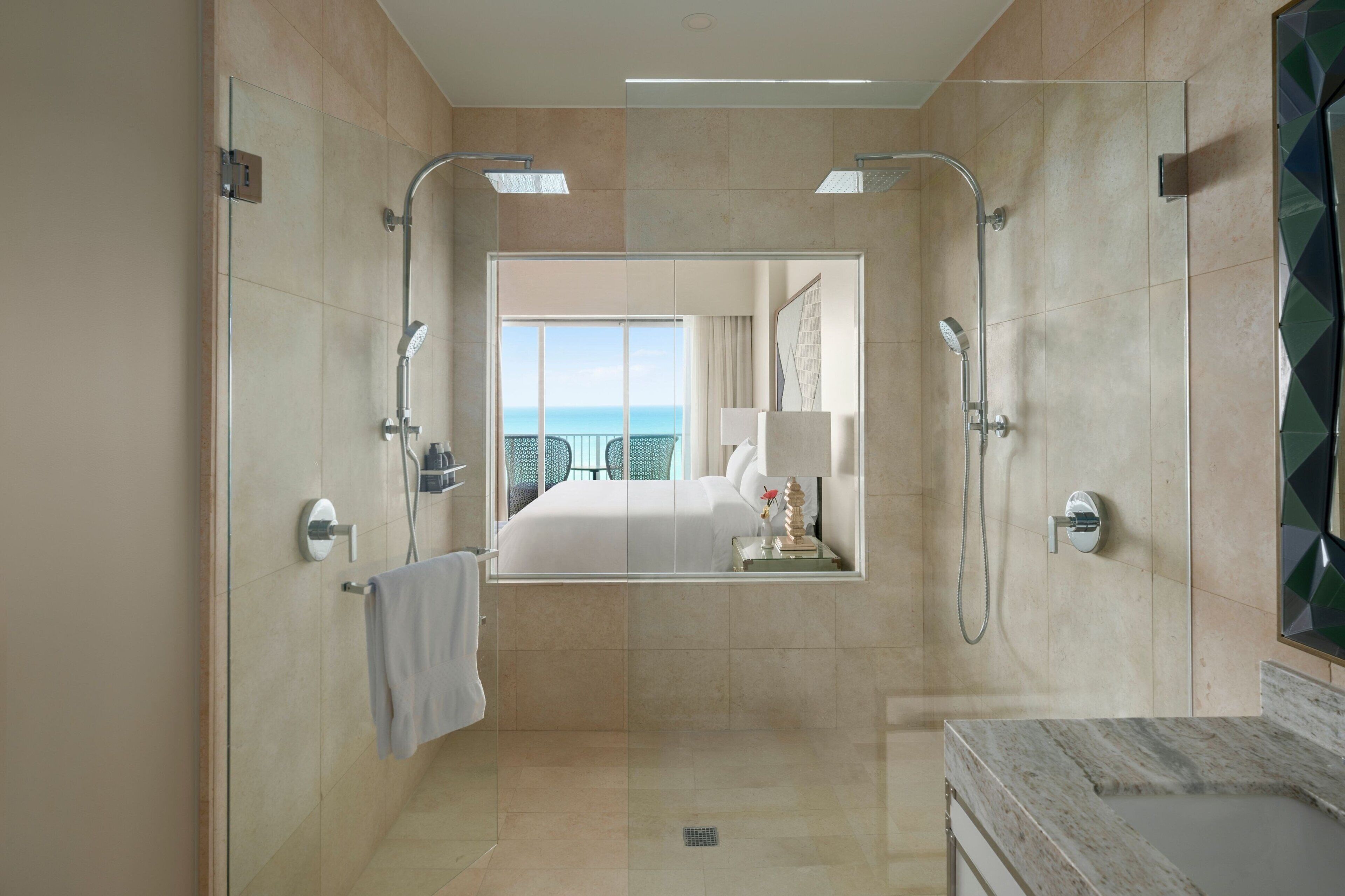 A modern bathroom with a glass-enclosed shower, a marble sink, and a large mirror reflecting a bedroom with a balcony view.