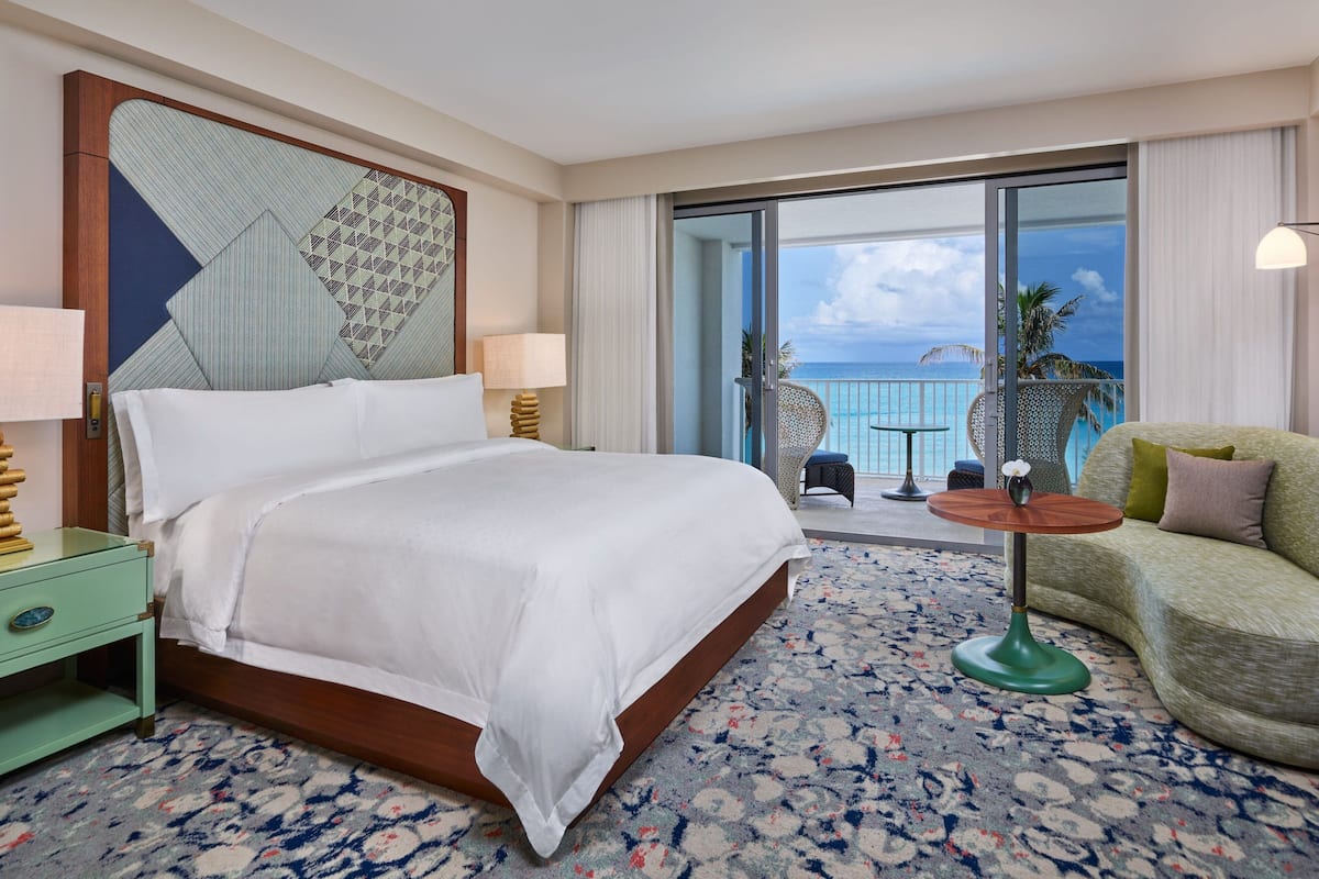 A hotel room with a large bed, a green armchair, a small table, and a balcony with a view of the ocean.