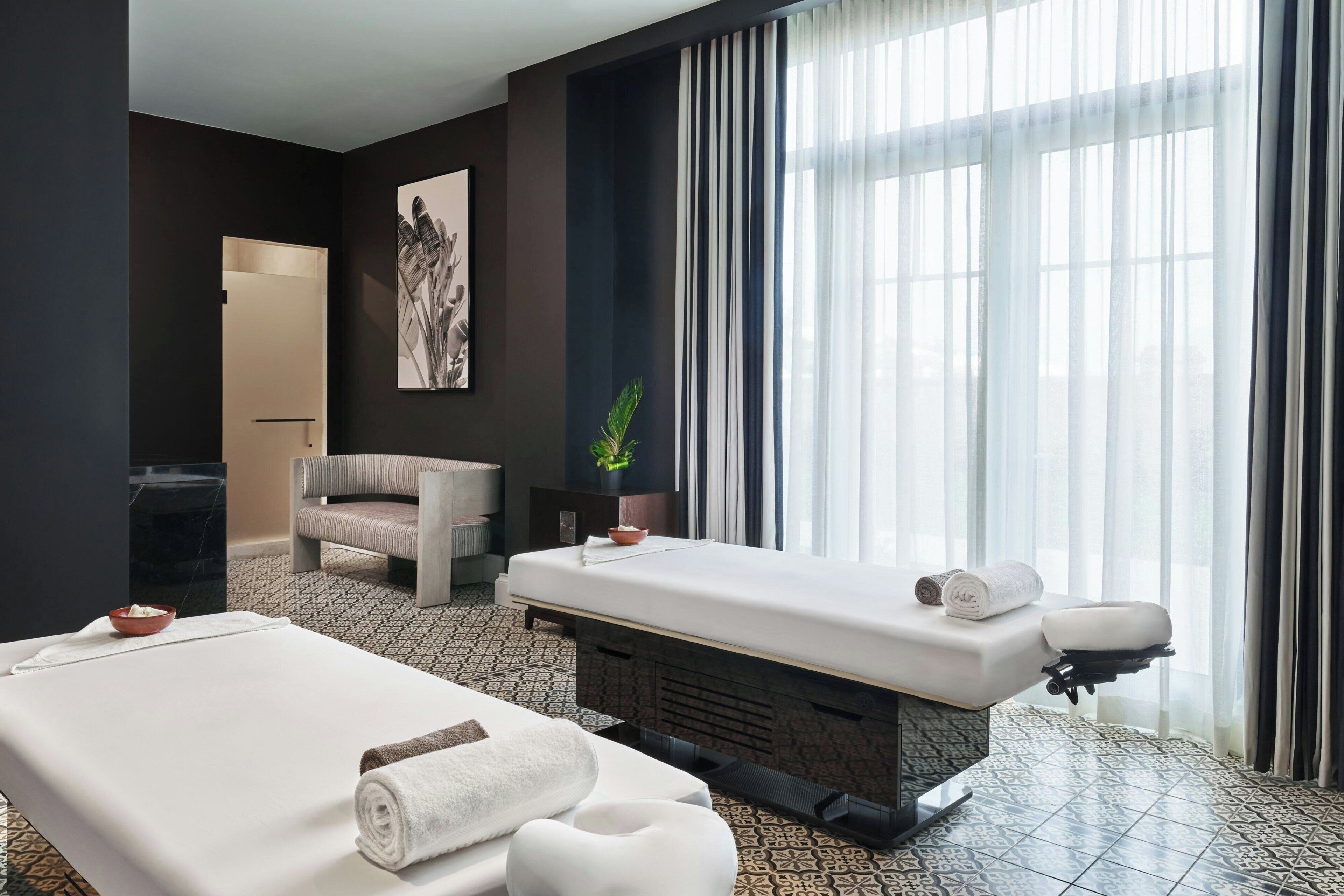 A modern hotel room with a massage table, a seating area, and a large window with sheer curtains.