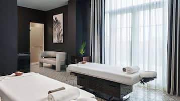 A modern hotel room with a massage table, a seating area, and a large window with sheer curtains.