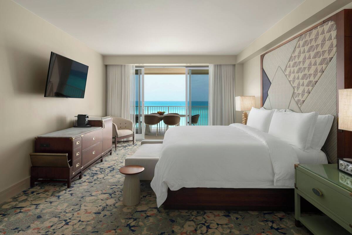 A hotel room with a large bed, a TV mounted on the wall, a desk, and a view of the ocean through the balcony doors.