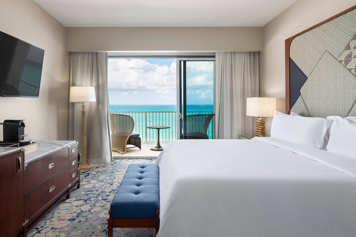 A hotel room with a large bed, a desk with drawers, a television, a balcony with a view of the ocean, and two chairs.