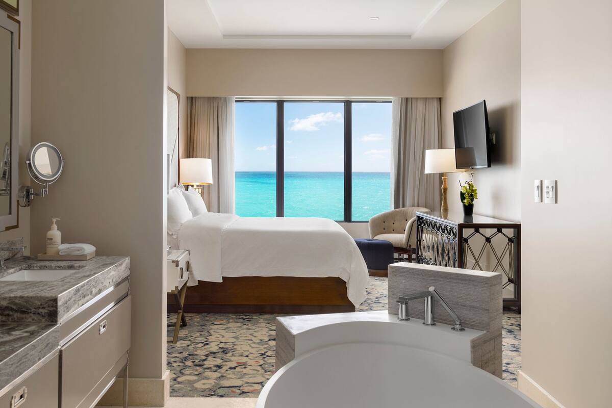 A hotel room with a large bed, a bathtub, a TV, and a view of the ocean.