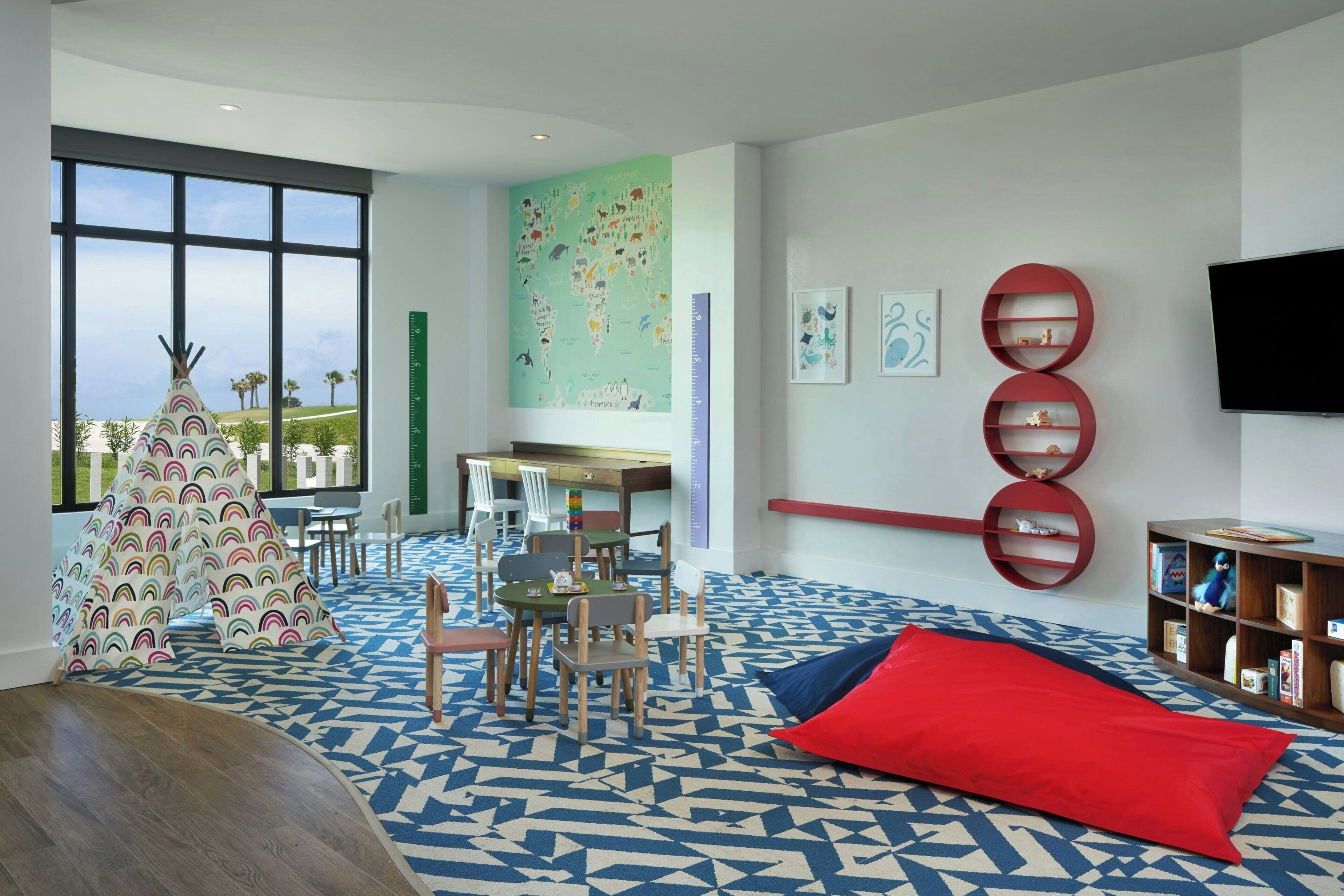 Un moderno salón infantil con un ventanal grande, una alfombra colorida y una estantería.