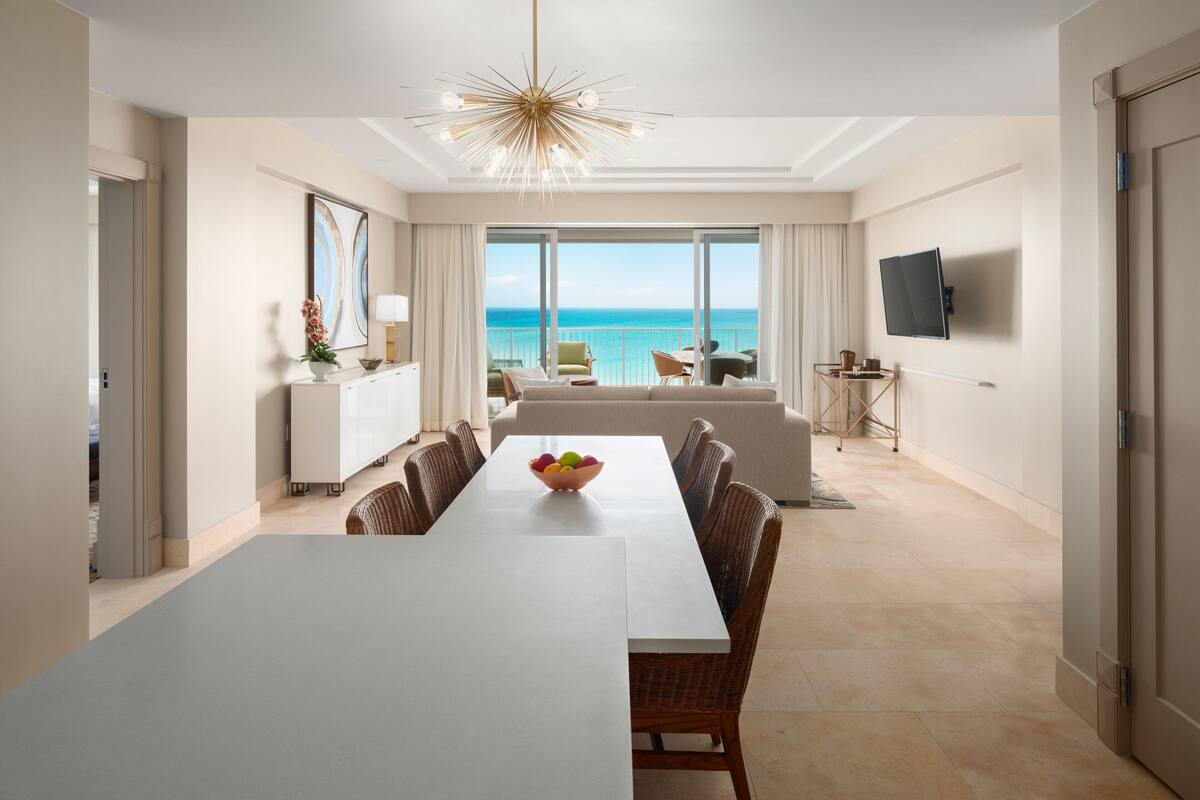 A modern hotel room with a large dining table, wicker chairs, a sofa, and a TV.