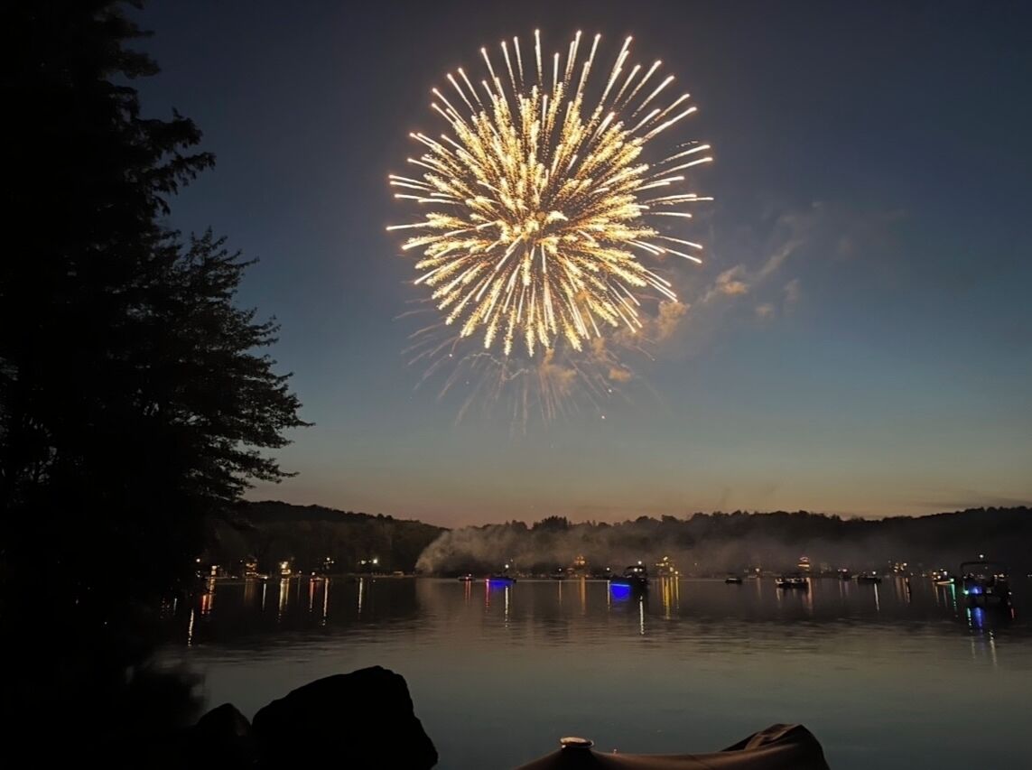 Un espectacle de focs artificials sobre un llac tranquil, amb vaixells i un turó boscós al fons.
