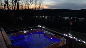 A hot tub with a wooden deck and lights, surrounded by trees and a view of the landscape. - Chalet w/ Lake & MT views, Hot Tub, Pet friendly,  <10mins to Lake & Golf (Wells)