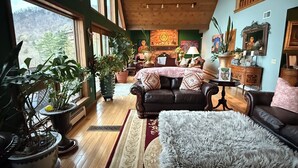 A spacious living room with a large window, wooden flooring, a leather sofa, and various potted plants. - Chalet w/ Lake & MT views, Hot Tub, Pet friendly,  <10mins to Lake & Golf (Wells)