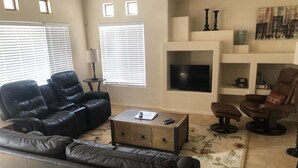 A living room with a black leather sofa, a coffee table, and a TV on a stand. There are two windows with blinds, a fireplace with a TV, and a small table with a lamp.