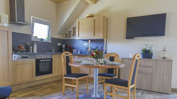 A modern kitchen with wooden cabinets, a built-in oven, a dining table with four chairs, and a flat-screen TV mounted on the wall. - Gîte au pré des oies (Ostheim)