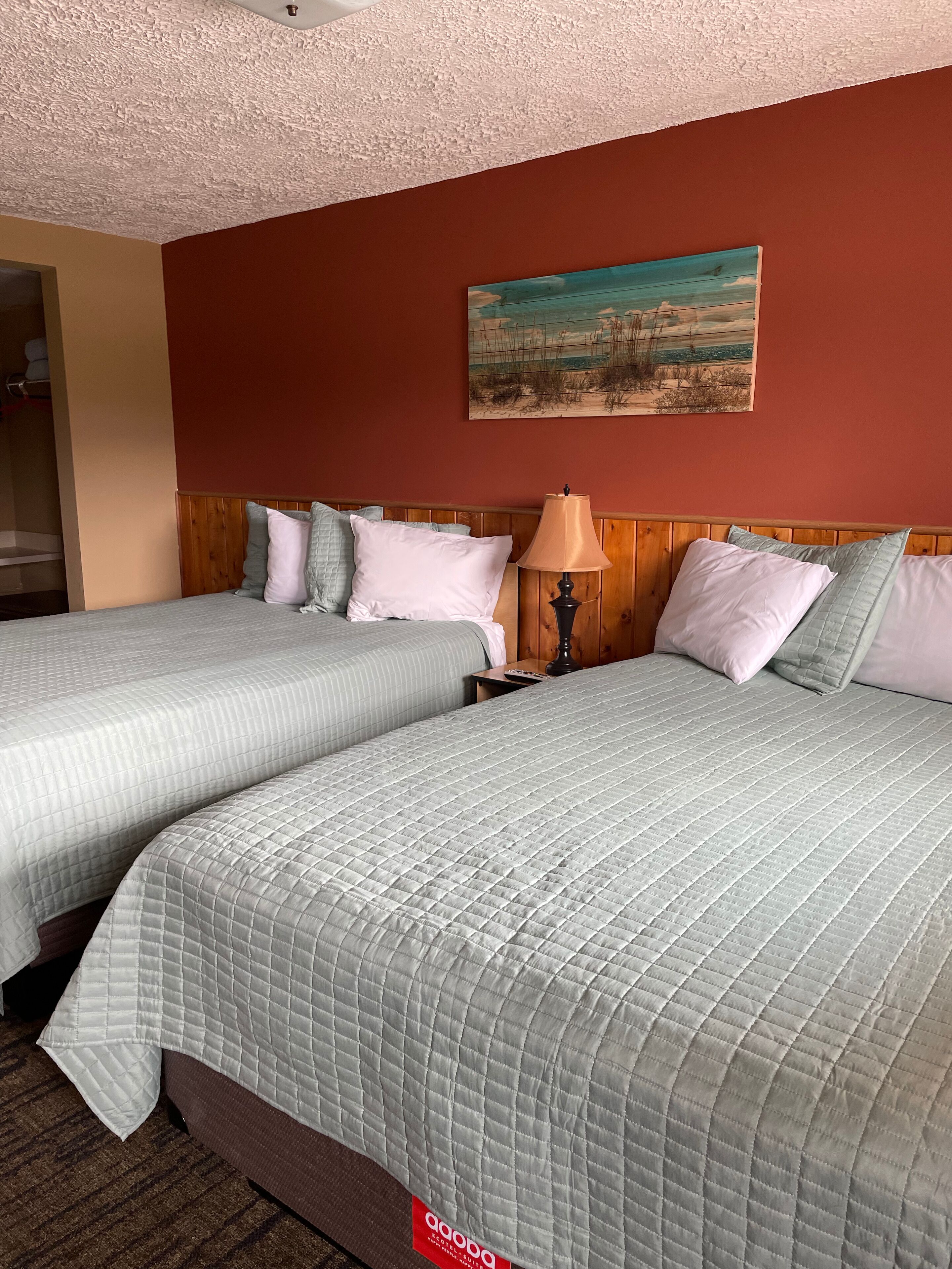 Quarto de hotel com duas camas, cabeceira de madeira, pintura na parede e abajur na mesa de cabeceira.