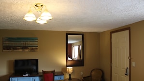 Et hotelværelse med fjernsyn, skrivebord, lampe, stol og dør.