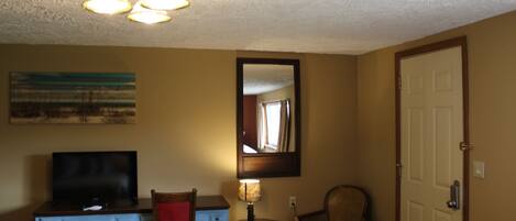 Een hotelkamer met een televisie, bureau, lamp, stoel en een deur.