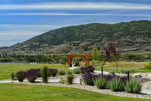 A park with a wooden pergola, landscaped garden beds, and a lake in the background. - Spacious Bordeaux Cottage with Golf Cart #191 (Osoyoos)