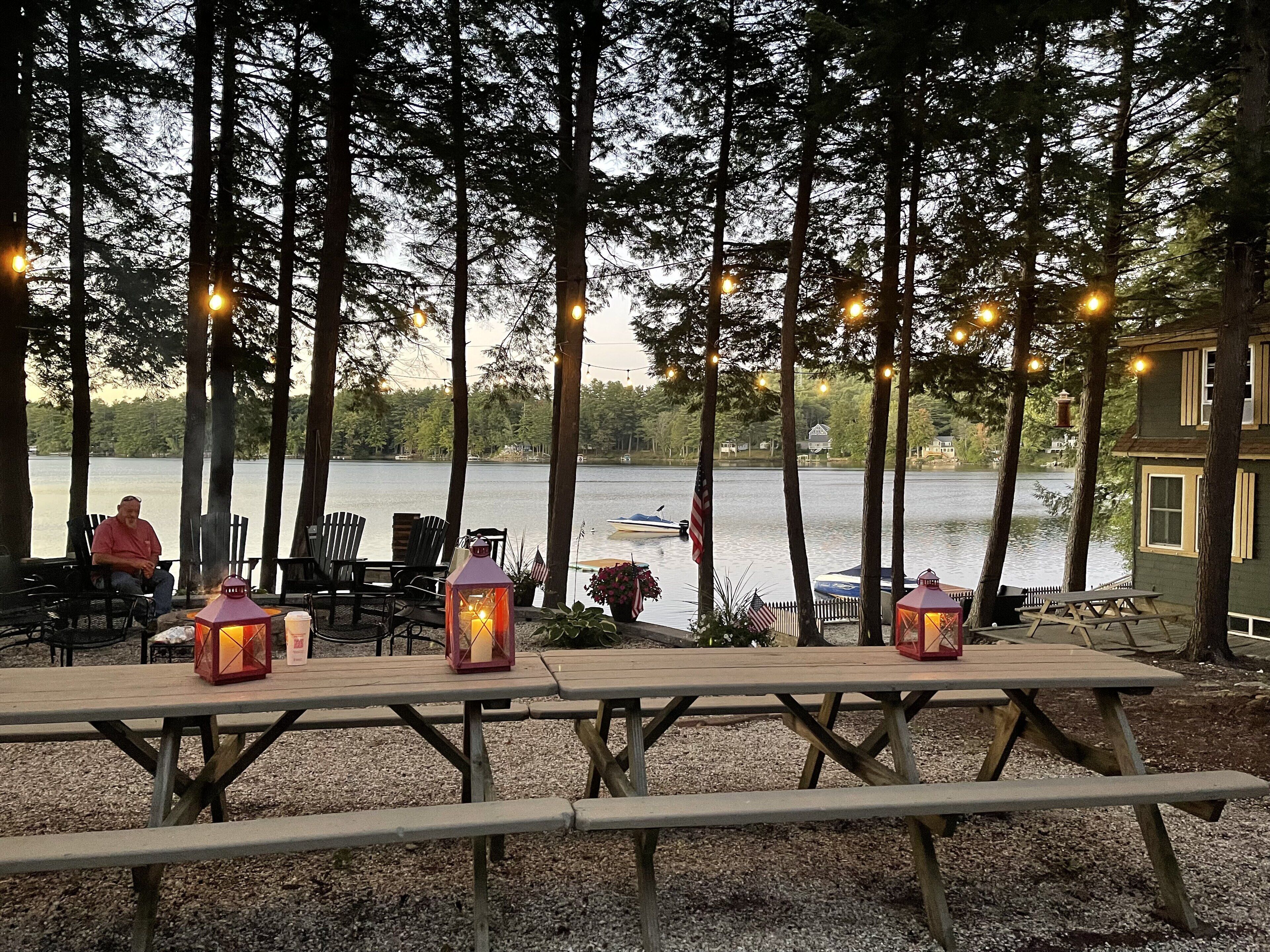 An outdoor dining area with wooden picnic tables and chairs, surrounded by trees and overlooking a lake.