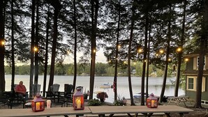 An outdoor dining area with wooden picnic tables and chairs, surrounded by trees and overlooking a lake. - Lake house, Dock, 3 beds/3 full baths. Pet friendly! (Shapleigh)