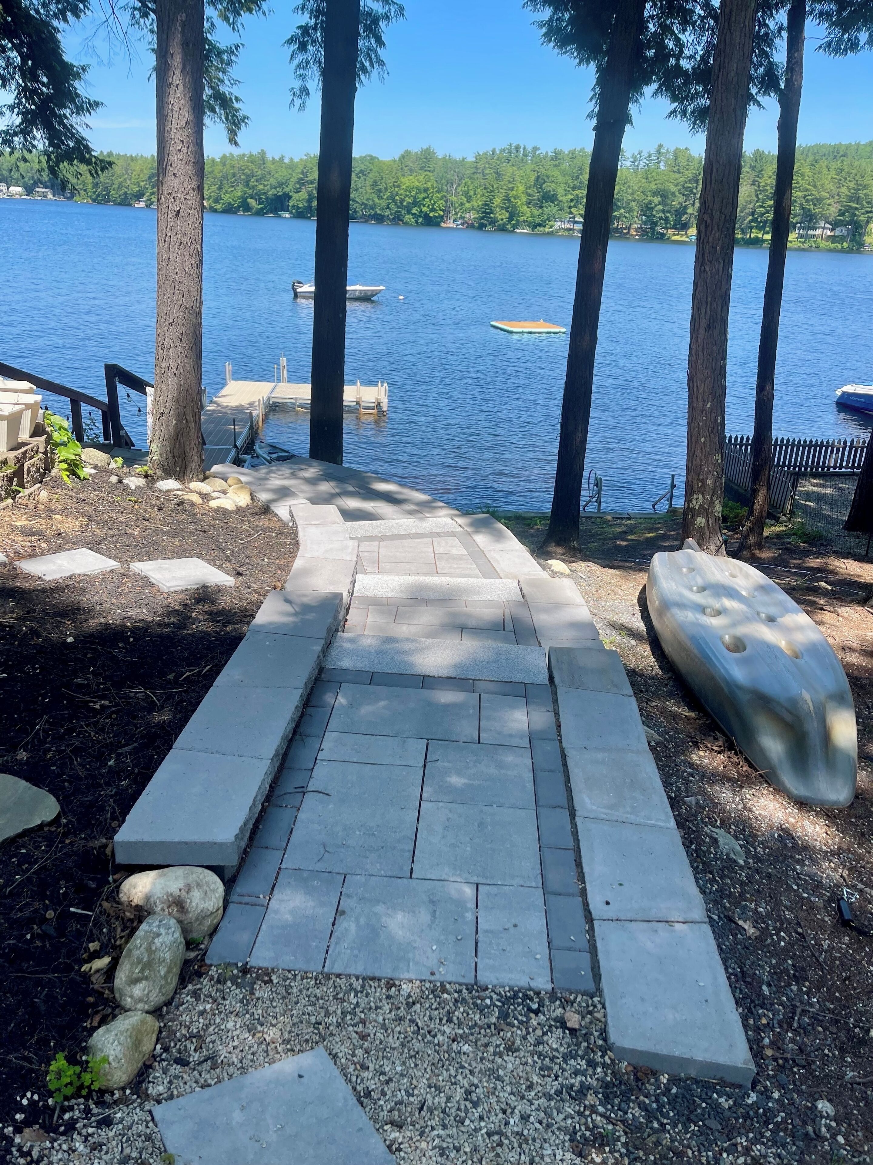 A stone pathway leading to a dock by a lake, surrounded by trees and a wooden fence.