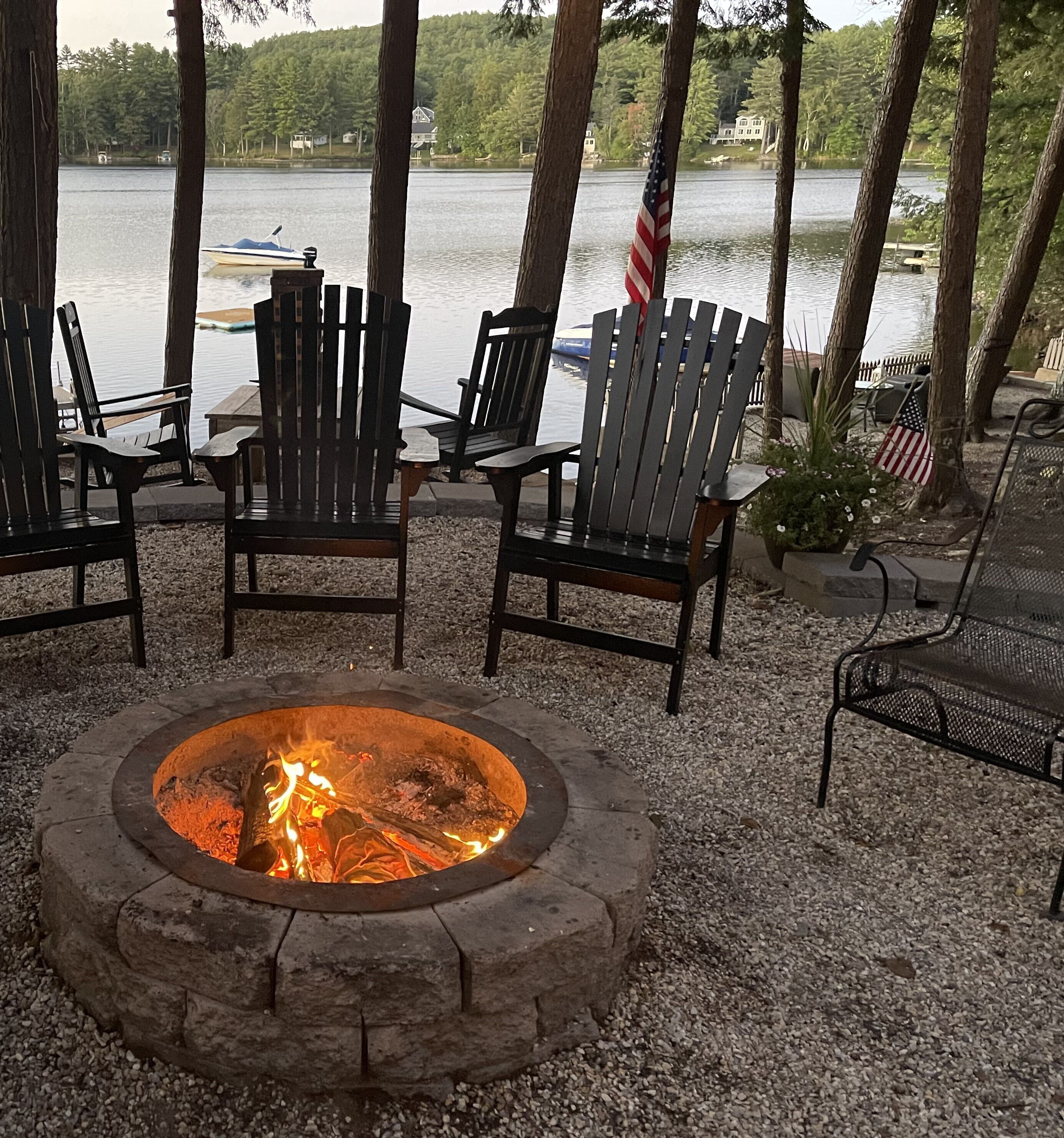 A fire pit surrounded by Adirondack chairs, overlooking a lake with boats and houses in the background.