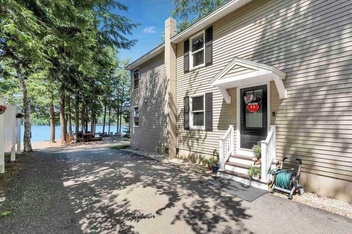 Une maison à deux étages avec un porche à l’avant, une pancarte de bienvenue et une vue sur un lac.