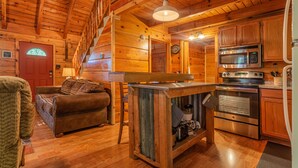 Un acogedor interior de cabaña con una escalera de madera, una sala de estar con sofá, una cocina con electrodomésticos modernos y una mesa de comedor de madera.