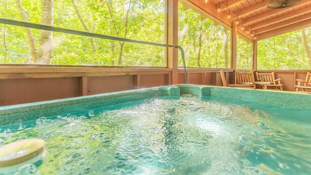 Un jacuzzi con terraza de madera y sillas, rodeado de grandes ventanales con vista al bosque.