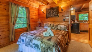 Un dormitorio de cabaña acogedor con interior de madera, una cama con colcha, una mesita de noche, una lámpara, un cuadro enmarcado en la pared y vistas al exterior a través de una ventana.