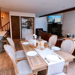 A dining area with a wooden table set for six, white chairs, and a television mounted on the wall.
