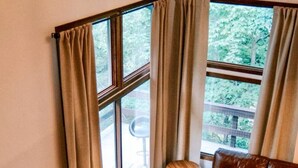 A cozy living room with a brown leather sofa, a wooden coffee table, and a small round table with a chair. Large windows with beige curtains allow natural light and a view of greenery outside. - Family Retreat with Sauna and Hot Tub (Tannersville)
