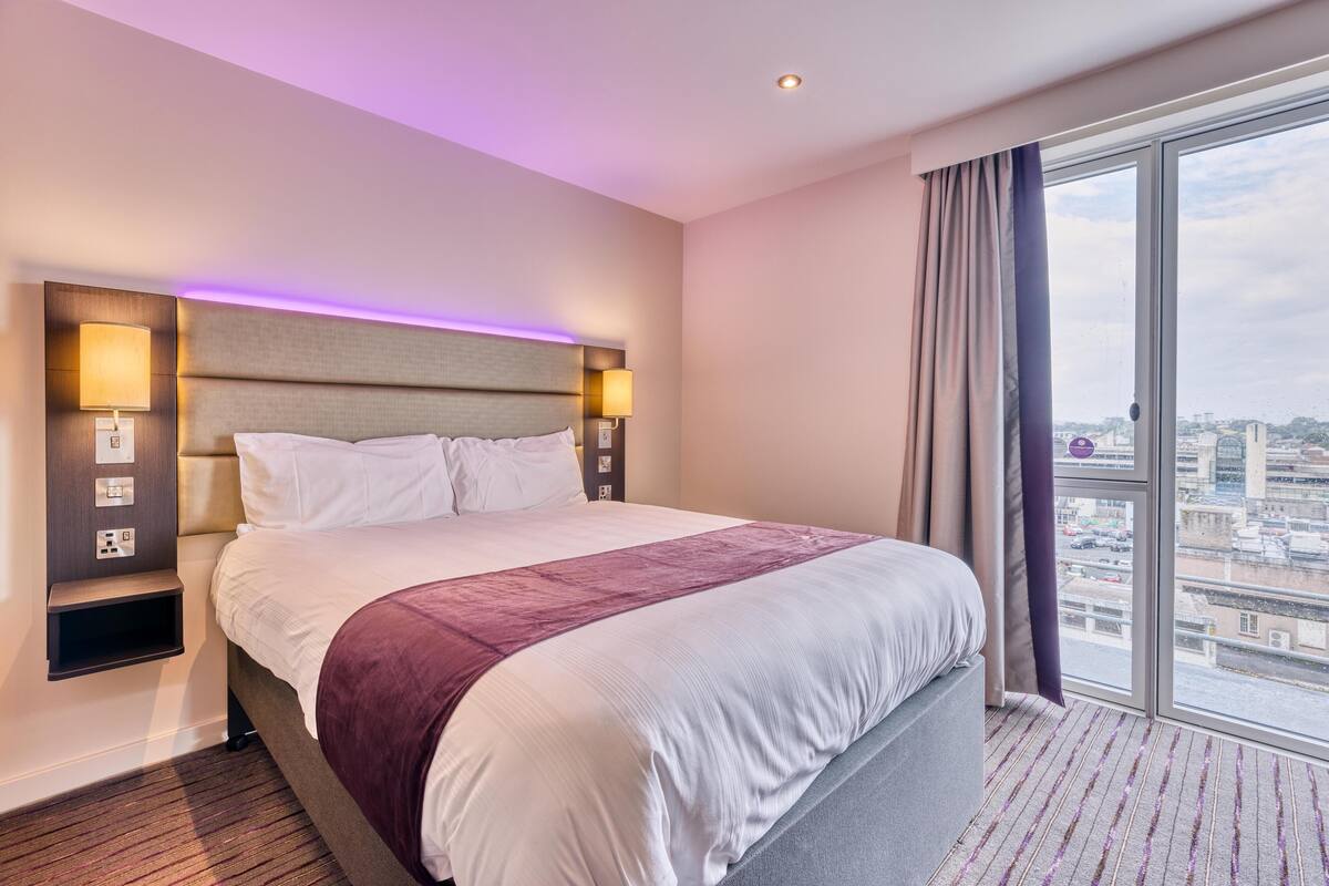 A hotel room with a large bed, a headboard with integrated lighting, a view of the city through large windows, and a built-in shelf with a lamp.