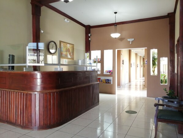 A reception area with a wooden reception desk, a clock, framed artwork, and a view into a hallway with a tiled floor and a potted plant.