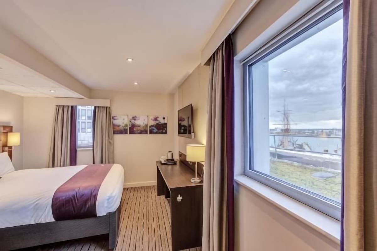 Habitación de hotel con cama, escritorio, silla, ventana con vista a un barco y pared con cuadros enmarcados.