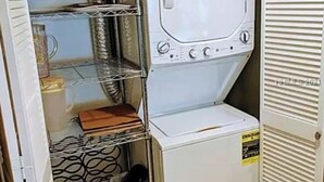 Kompakti pyykinhuoltotila, jossa on etuaukosta täytettävä pesukone, metallinen hyllykkö ja roskakori.
