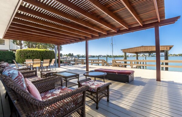 Een terras buiten met rieten meubilair, een houten pergola en uitzicht op het water.