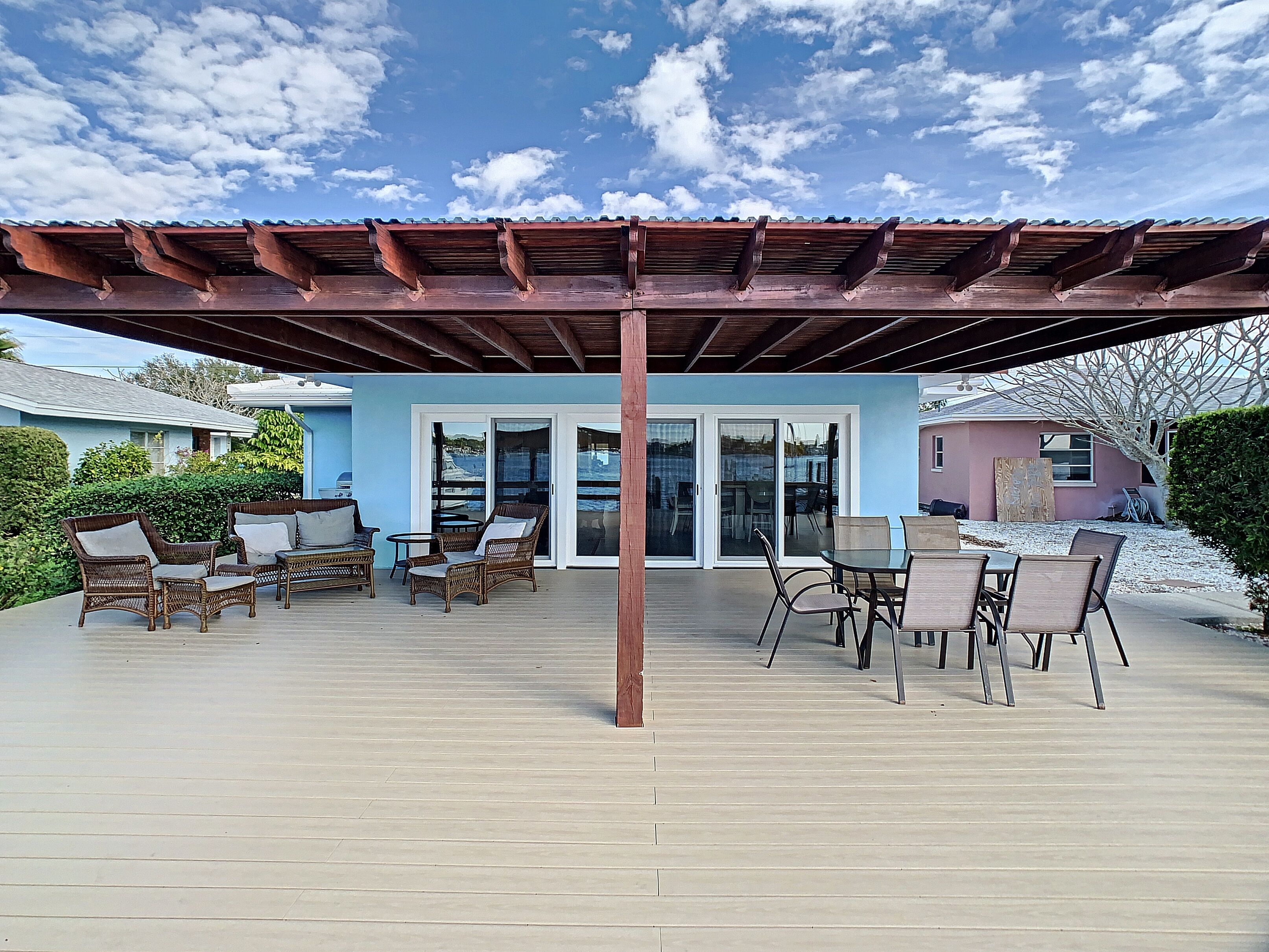 En overdækket terrasse med fletningsmøbler, et pergola og udsigt til et hus med blå facade.
