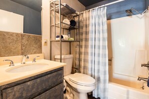 Un baño con un lavabo blanco, un inodoro, una ducha con cortina y un estante con toallas y artículos de aseo.