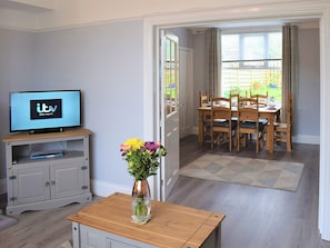 A living room with a television, a dining table with chairs, a vase with flowers, and a door leading to a garden.