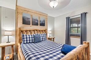 Una cama de madera con colcha a cuadros, dos almohadas y cabecera con obra de arte. Hay dos mesitas de noche con lámparas.