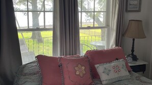 A bedroom with a bed, pillows, a nightstand, a lamp, and a view of a garden through large windows.