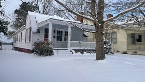 A snow-covered house with a porch, a small bush, and a tree in front.