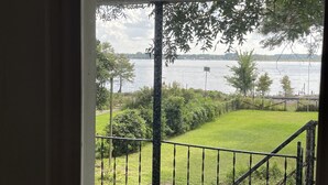 A view from a window overlooking a grassy area with a metal railing, a body of water, and trees in the background.