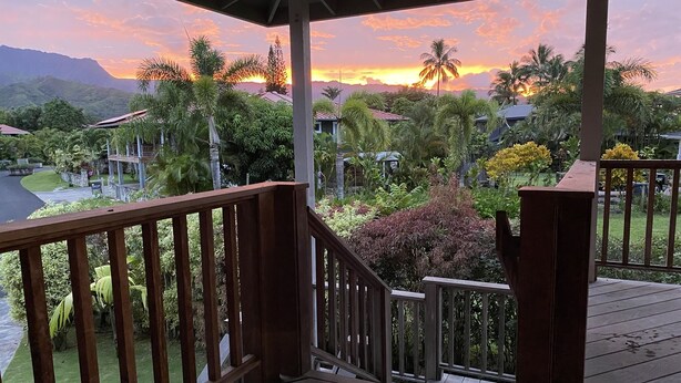 A wooden deck with a view of a sunset over a tropical landscape with palm trees and houses. - Heavenly Hanalei Home in Tropical Paradise (Hanalei)