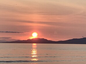 Auringonlasku tyynen vesistön yllä, ja kauempana horisontissa siintää vuoriston siluetti.