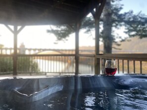 Un jacuzzi con una copa de vino tinto en una terraza de madera con vistas a un cuerpo de agua.