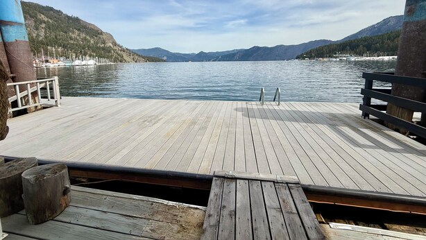 A wooden deck with a metal railing, overlooking a calm lake with mountains in the background. - The Bear Den (Bayview)