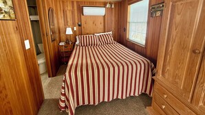 A wooden-paneled bedroom with a bed, bedside table, lamp, and a window with blinds. - The Bear Den (Bayview)