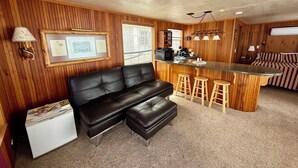 A cozy cabin interior with a black leather sofa, wooden bar stools, a small kitchenette, and a striped couch. - The Bear Den (Bayview)