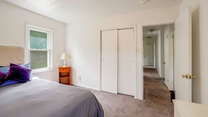 Un dormitorio con una cama, una mesita de noche, una ventana y una puerta que da a un pasillo.