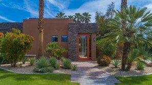 A modern building with a stone facade, a glass door entrance, and a well-maintained garden with palm trees and flowering plants.