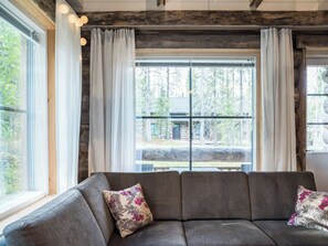 Una habitación con sofá gris, cortinas blancas y vista a una cabaña de madera a través de amplias ventanas.
