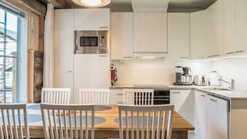 A modern kitchen with white cabinetry, a built-in oven, a microwave, and a coffee maker. There is a dining table with chairs and a hanging light fixture above.